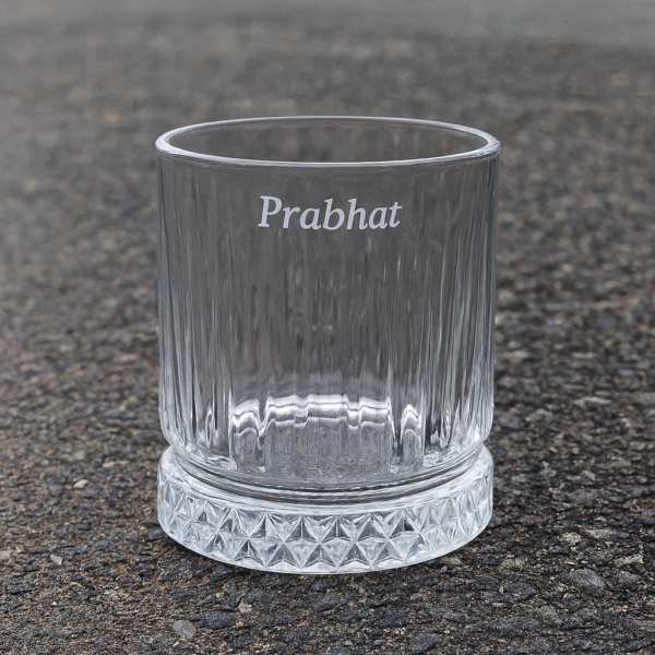 Personalized engraved whiskey glass with name 'Prabhat' on the side and bottom message 'I Promise to Always Be by Your Side', displayed outdoors on grass