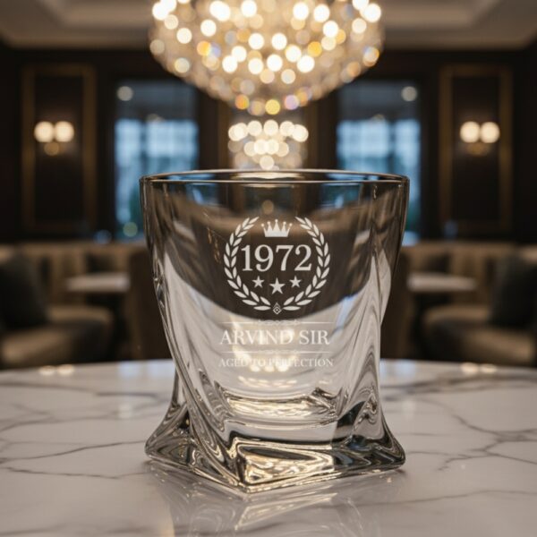 A personalized crystal-clear whiskey glass with deep engraving featuring the year "1972", a laurel wreath, and the name "Arvind Sir" above the phrase "Aged to Perfection". The glass has a twisted base and is placed on a dark stone surface beside an ice cube. Warm ambient lighting in the background highlights the clarity and premium craftsmanship of the glass.
