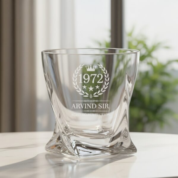A personalized crystal-clear whiskey glass with deep engraving featuring the year "1972", a laurel wreath, and the name "Arvind Sir" above the phrase "Aged to Perfection". The glass has a twisted base and is placed on a dark stone surface beside an ice cube. Warm ambient lighting in the background highlights the clarity and premium craftsmanship of the glass.