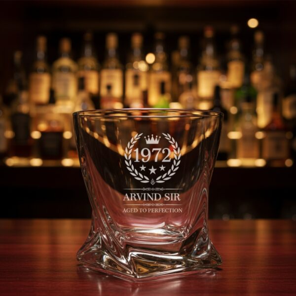 A personalized crystal-clear whiskey glass with deep engraving featuring the year "1972", a laurel wreath, and the name "Arvind Sir" above the phrase "Aged to Perfection". The glass has a twisted base and is placed on a dark stone surface beside an ice cube. Warm ambient lighting in the background highlights the clarity and premium craftsmanship of the glass.