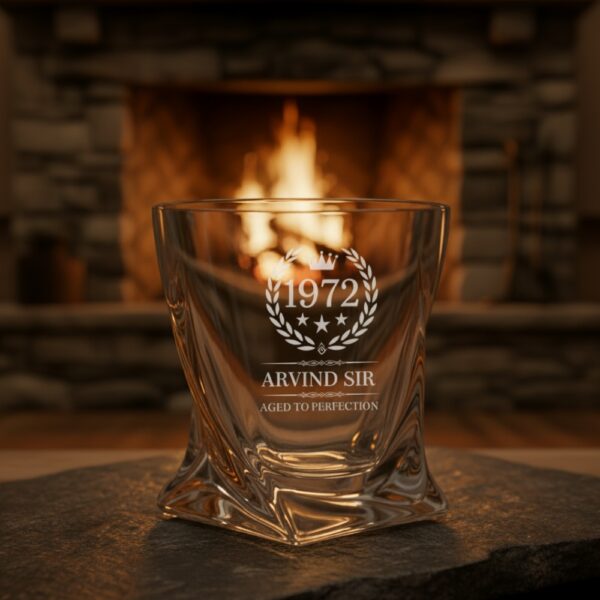 A personalized crystal-clear whiskey glass with deep engraving featuring the year "1972", a laurel wreath, and the name "Arvind Sir" above the phrase "Aged to Perfection". The glass has a twisted base and is placed on a dark stone surface beside an ice cube. Warm ambient lighting in the background highlights the clarity and premium craftsmanship of the glass.