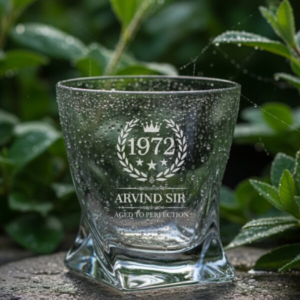 A personalized crystal-clear whiskey glass with deep engraving featuring the year "1972", a laurel wreath, and the name "Arvind Sir" above the phrase "Aged to Perfection". The glass has a twisted base and is placed on a dark stone surface beside an ice cube. Warm ambient lighting in the background highlights the clarity and premium craftsmanship of the glass.
