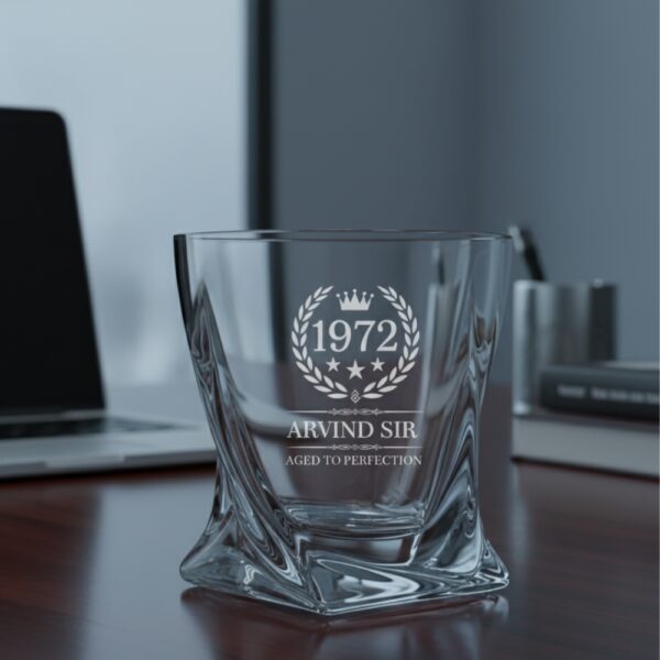 A personalized crystal-clear whiskey glass with deep engraving featuring the year "1972", a laurel wreath, and the name "Arvind Sir" above the phrase "Aged to Perfection". The glass has a twisted base and is placed on a dark stone surface beside an ice cube. Warm ambient lighting in the background highlights the clarity and premium craftsmanship of the glass.