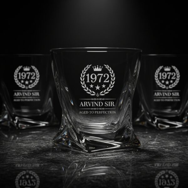 A personalized crystal-clear whiskey glass with deep engraving featuring the year "1972", a laurel wreath, and the name "Arvind Sir" above the phrase "Aged to Perfection". The glass has a twisted base and is placed on a dark stone surface beside an ice cube. Warm ambient lighting in the background highlights the clarity and premium craftsmanship of the glass.