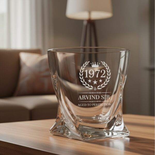 A personalized crystal-clear whiskey glass with deep engraving featuring the year "1972", a laurel wreath, and the name "Arvind Sir" above the phrase "Aged to Perfection". The glass has a twisted base and is placed on a dark stone surface beside an ice cube. Warm ambient lighting in the background highlights the clarity and premium craftsmanship of the glass.