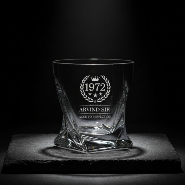 A personalized crystal-clear whiskey glass with deep engraving featuring the year "1972", a laurel wreath, and the name "Arvind Sir" above the phrase "Aged to Perfection". The glass has a twisted base and is placed on a dark stone surface beside an ice cube. Warm ambient lighting in the background highlights the clarity and premium craftsmanship of the glass.