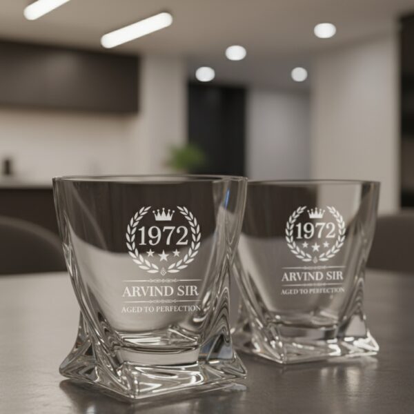 A personalized crystal-clear whiskey glass with deep engraving featuring the year "1972", a laurel wreath, and the name "Arvind Sir" above the phrase "Aged to Perfection". The glass has a twisted base and is placed on a dark stone surface beside an ice cube. Warm ambient lighting in the background highlights the clarity and premium craftsmanship of the glass.