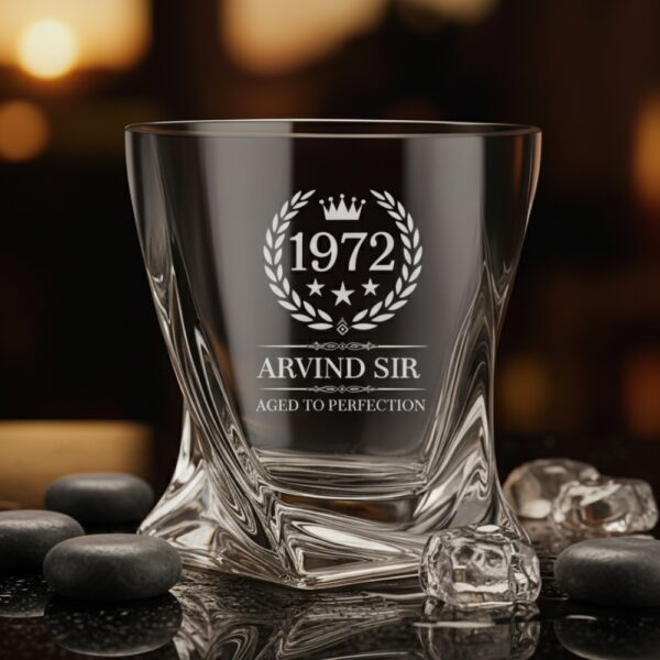 A personalized crystal-clear whiskey glass with deep engraving featuring the year "1972", a laurel wreath, and the name "Arvind Sir" above the phrase "Aged to Perfection". The glass has a twisted base and is placed on a dark stone surface beside an ice cube. Warm ambient lighting in the background highlights the clarity and premium craftsmanship of the glass.